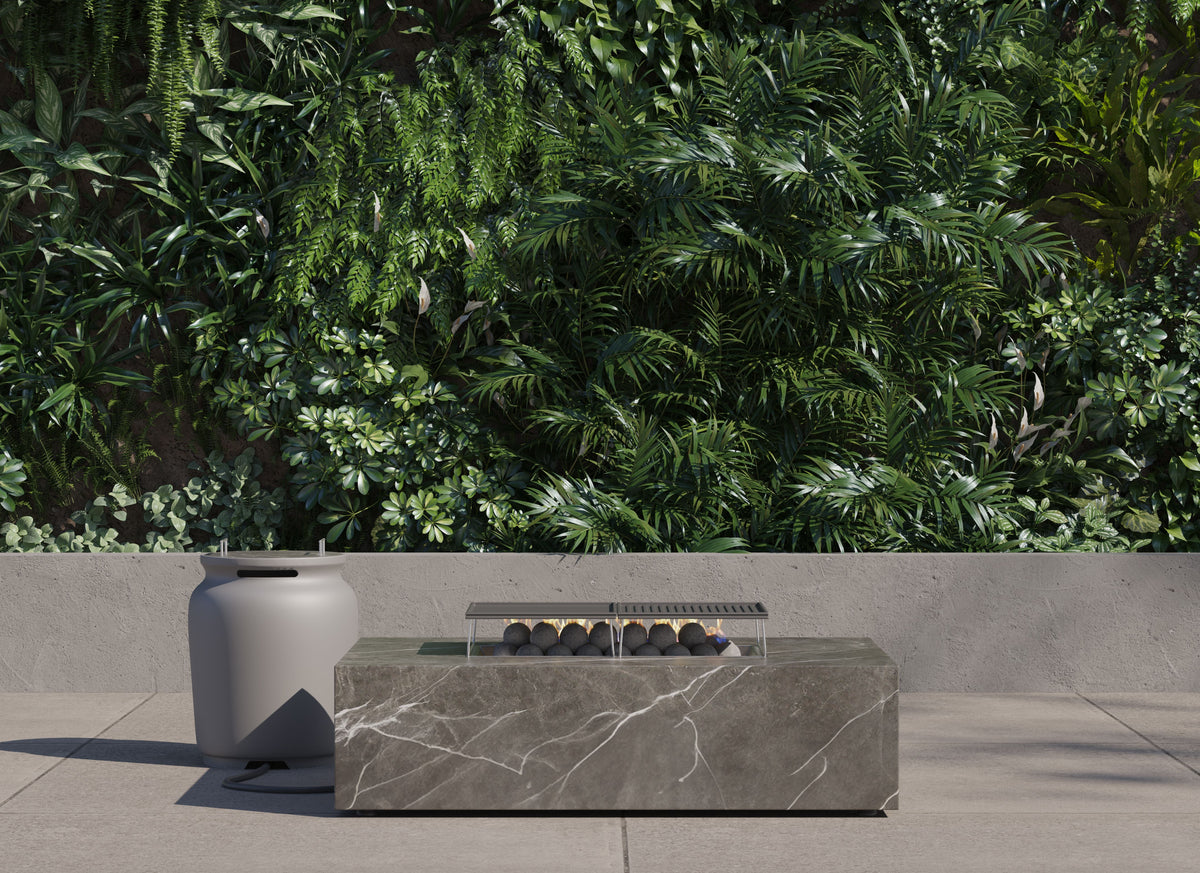 Rectangular OuterStone® fire pit table with a stone finish, cast-iron griddles, and gray propane tank cover beside lush greenery.