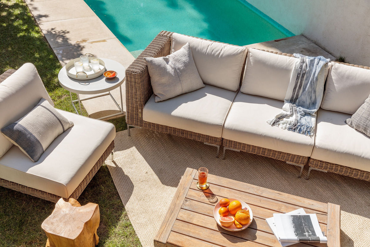 Handwoven brown wicker loveseat with cream cushions, paired with a wooden table and decorative items, near a pool.