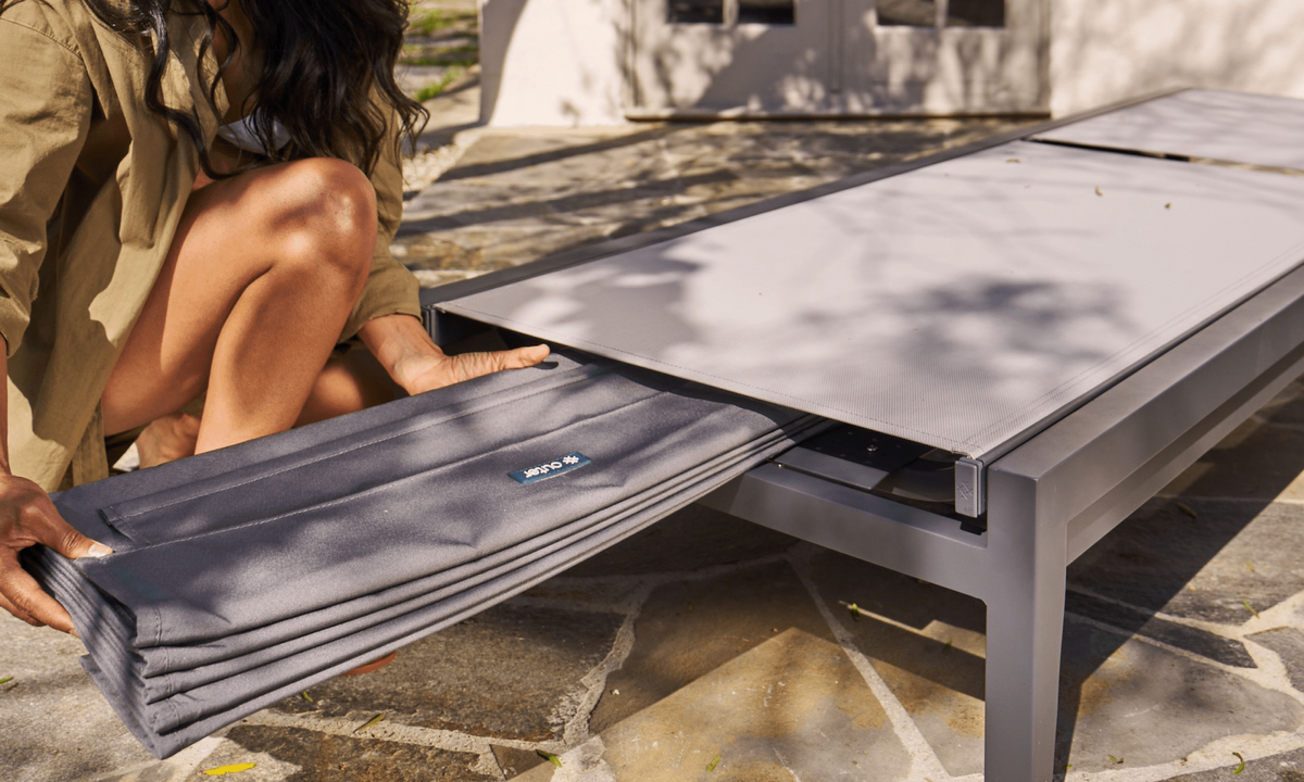 Charcoal aluminum chaise lounge with adjustable backrest, integrated cover, and a person unfolding the optional cushion.