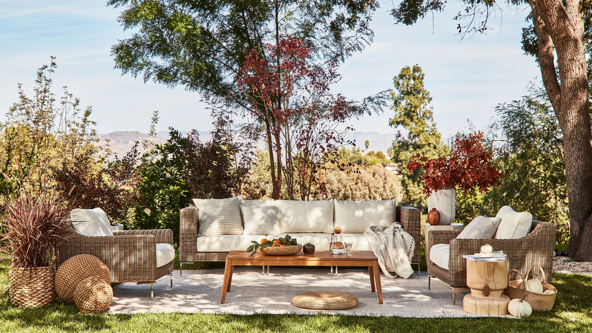 Handwoven brown wicker loveseat with cream cushions, arranged with two corner chairs, surrounded by greenery and decor.