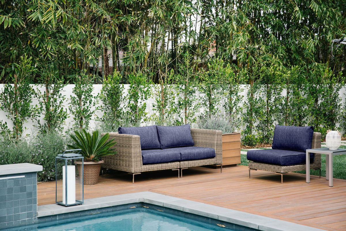Brown wicker outdoor loveseat with thick navy cushions, supported by polished stainless steel legs, set by a poolside.