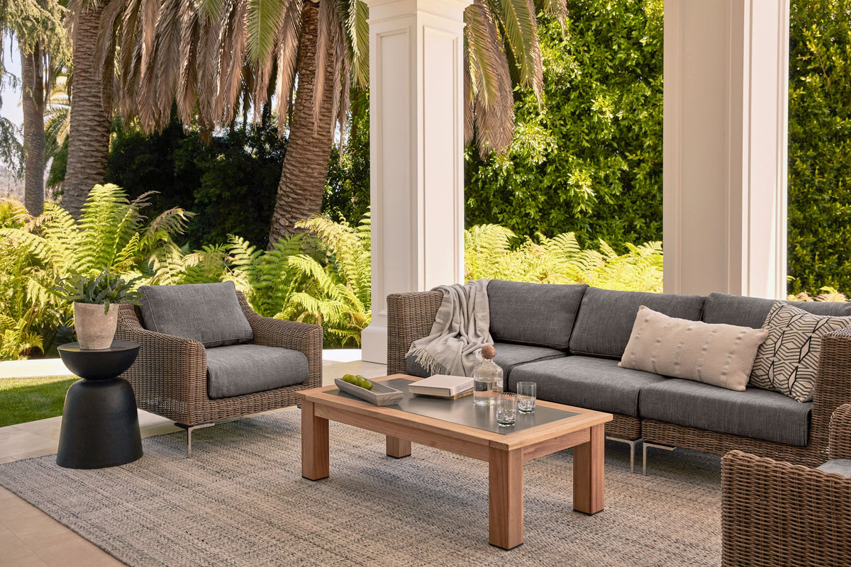 Handwoven brown wicker loveseat with gray cushions, modular design, and polished stainless steel legs, on a patio setting.