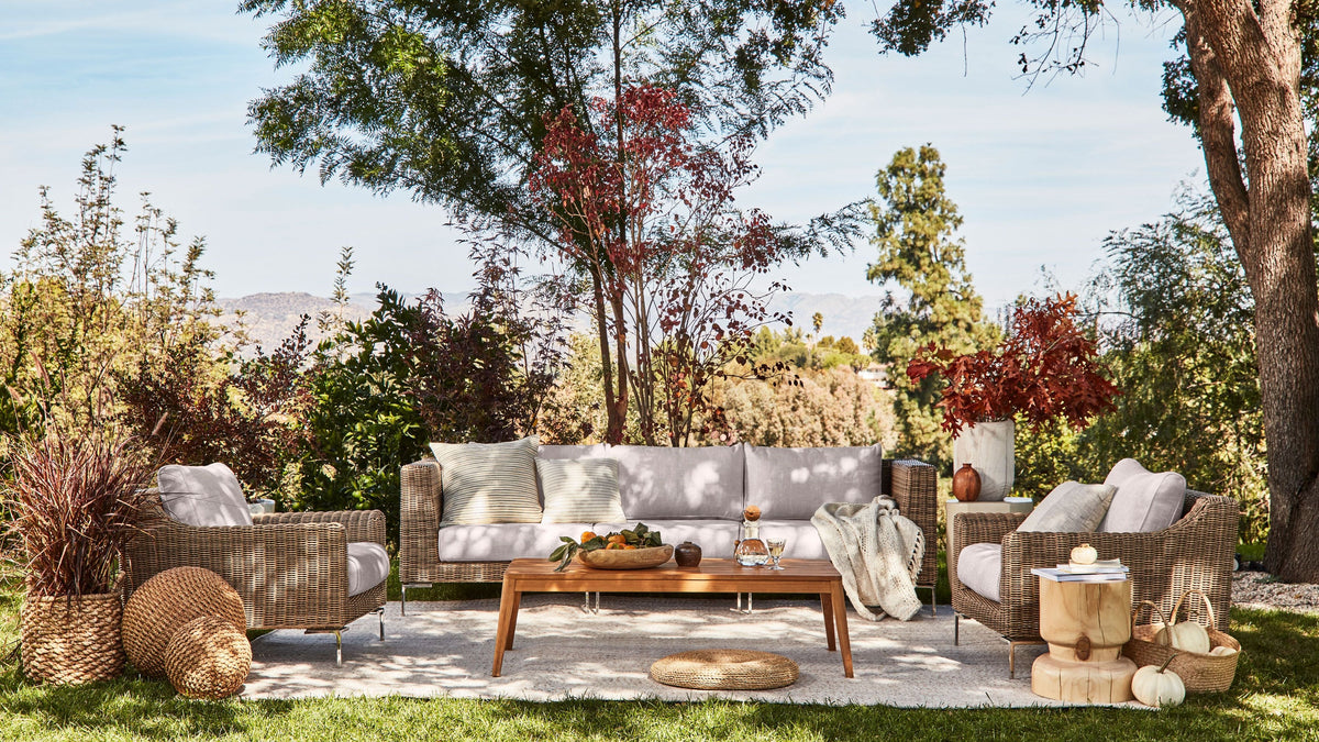 Handwoven brown wicker outdoor sofa set with two armchairs, gray cushions, and a wooden coffee table in a garden setting.