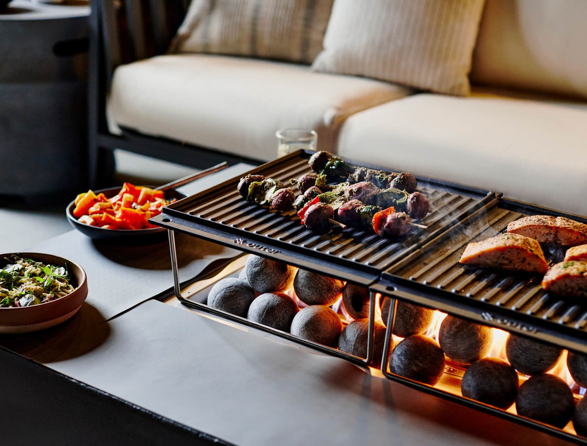 Rectangular OuterStone® fire pit cooking set featuring grilled meat and vegetables on cast-iron griddles, surrounded by decorative ceramic spheres.