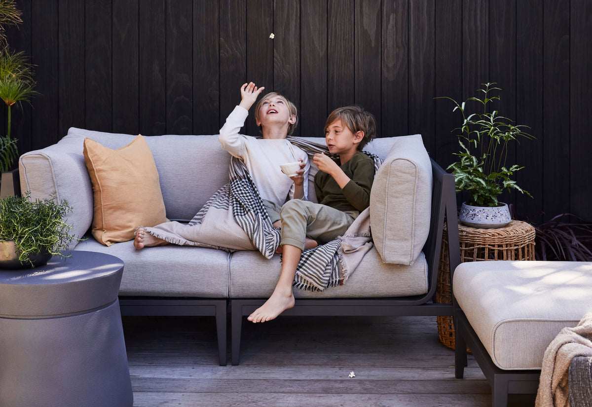 Charcoal aluminum three-seat outdoor sofa with light gray cushions, featuring two kids enjoying snacks with a throw blanket.