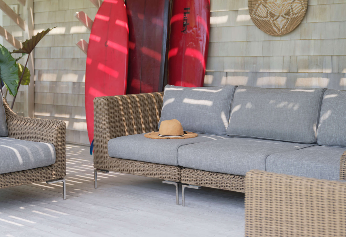 Handwoven brown wicker outdoor loveseat with gray cushions, polished stainless steel legs, and a straw hat on top.