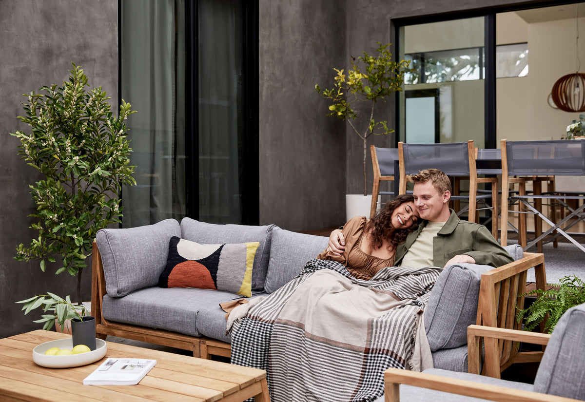 Three-seat outdoor sofa made of FSC-certified teak with layered gray cushions, featuring a cozy blanket and decorative pillows.